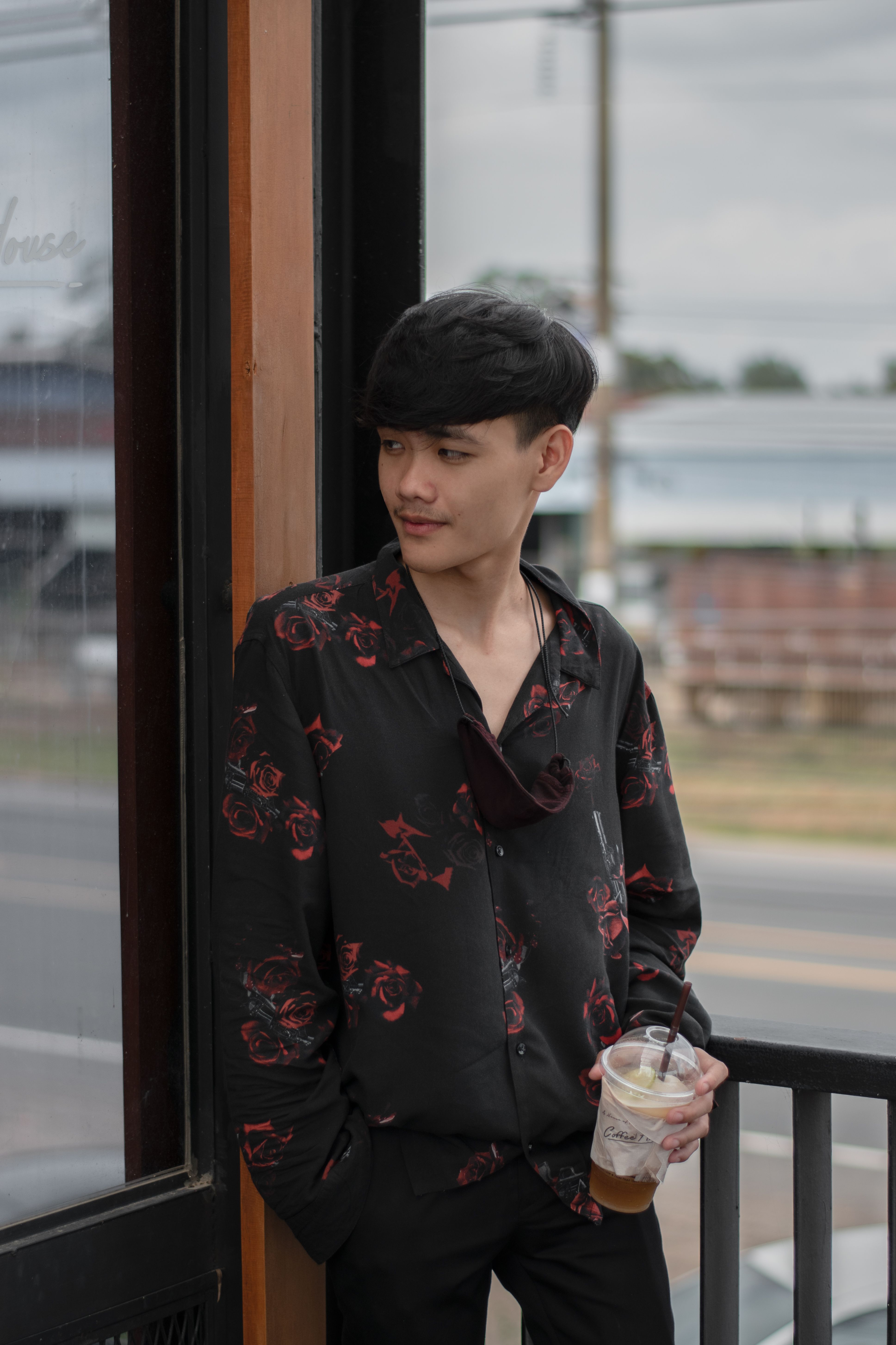 Young man in floral shirt looks away while holding iced coffee outdoors.