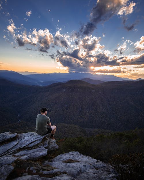 High angle back view of unrecognizable male explorer sitting on edge of rocky cliff and admiring picturesque scenery of highlands