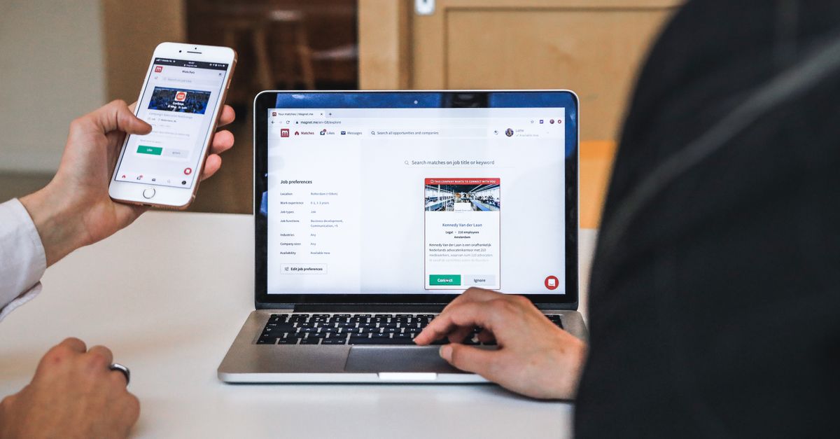 Hands using smartphone and laptop for digital browsing in an office setting, showcasing remote work culture.