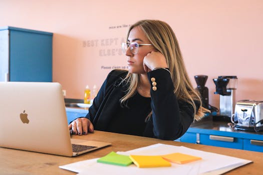 Blond woman in black blazer with glasses, working on laptop in modern office.