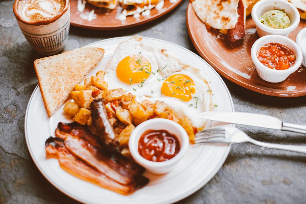 Two eggs with bacon, home fries and buttered toast on a diner plate beside a mug of coffee
