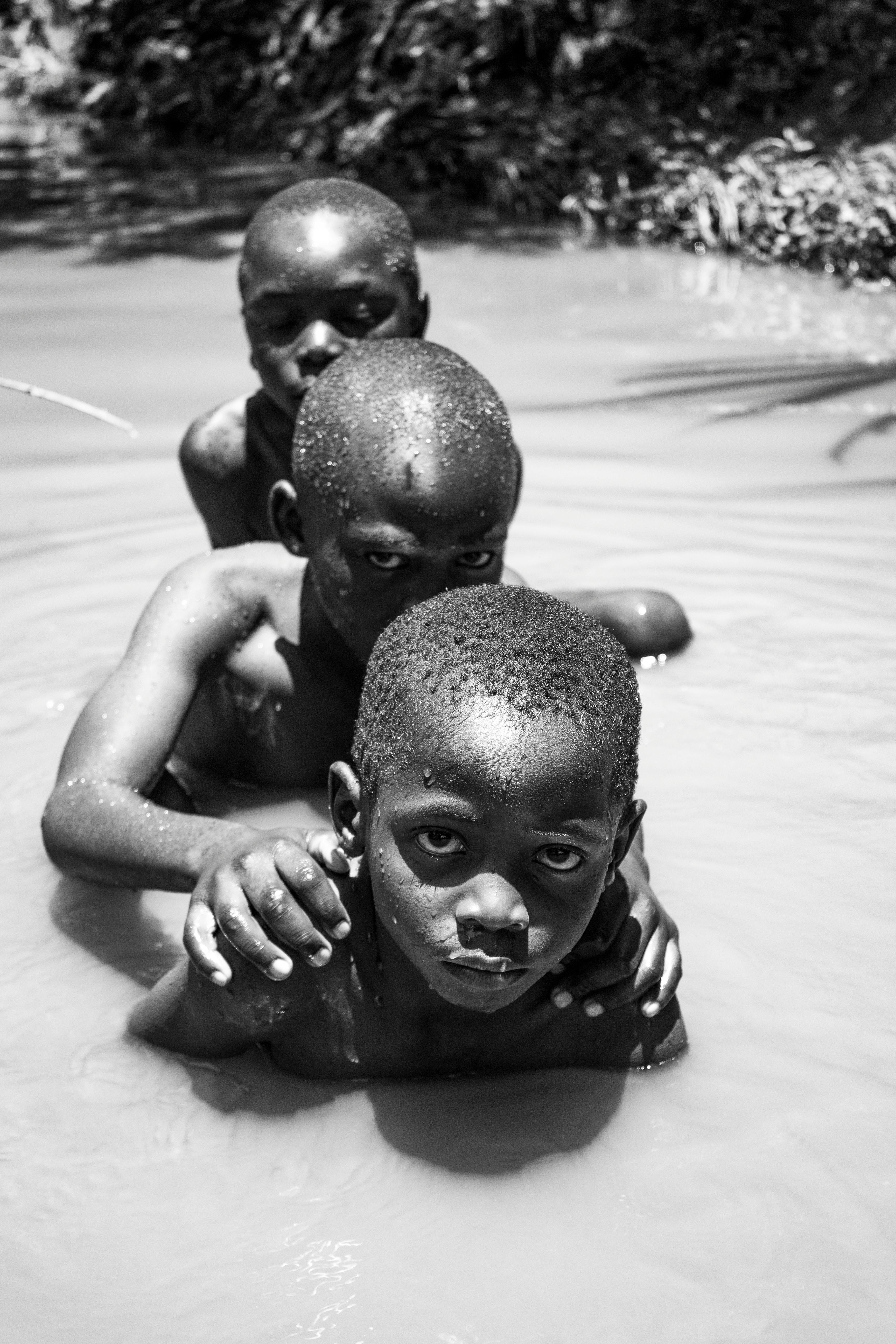 Three young boys swimming in muddy water in Kilgoris, Kenya, in a black and white photo.