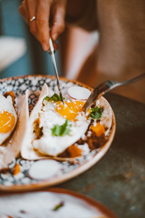 Free A person enjoys cutting into savory tacos topped with sunny side up eggs at a rustic table. Stock Photo