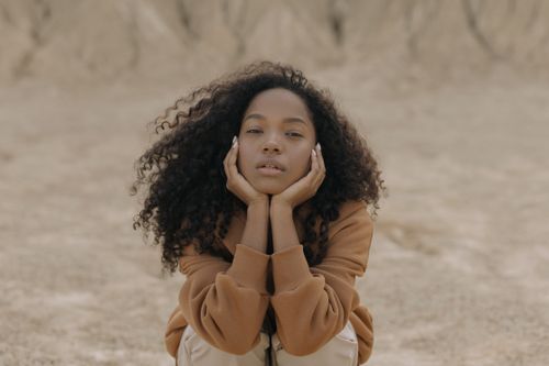 Free Portrait of a woman in a brown sweater sitting in a sandy landscape with hands on cheeks. Stock Photo
