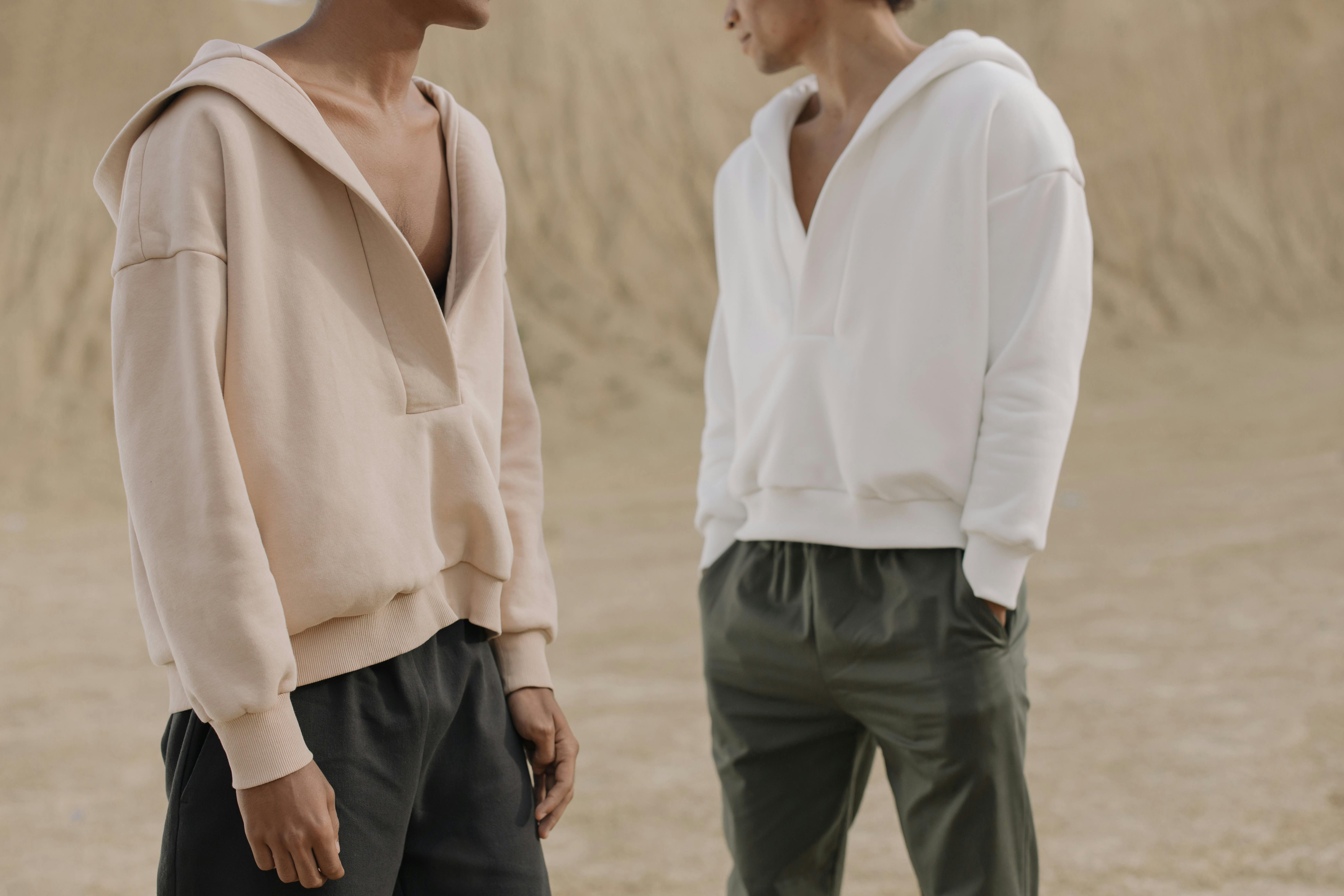 Two models wearing minimalist hoodies in a neutral outdoor setting. Modern fashion style captured.