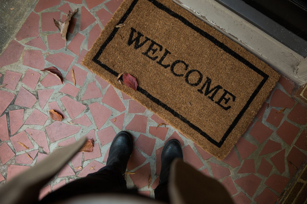 Top-down view of a coir ‘WELCOME’ doormat on a tiled front porch with winter boots at the door.