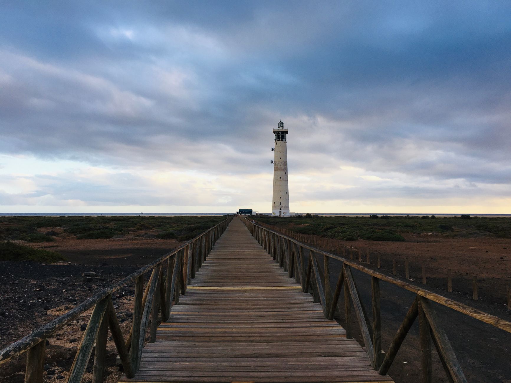 A picturesque wooden walkway leading to Faro de Morro Jable lighthouse under a cloudy sky in Spain.