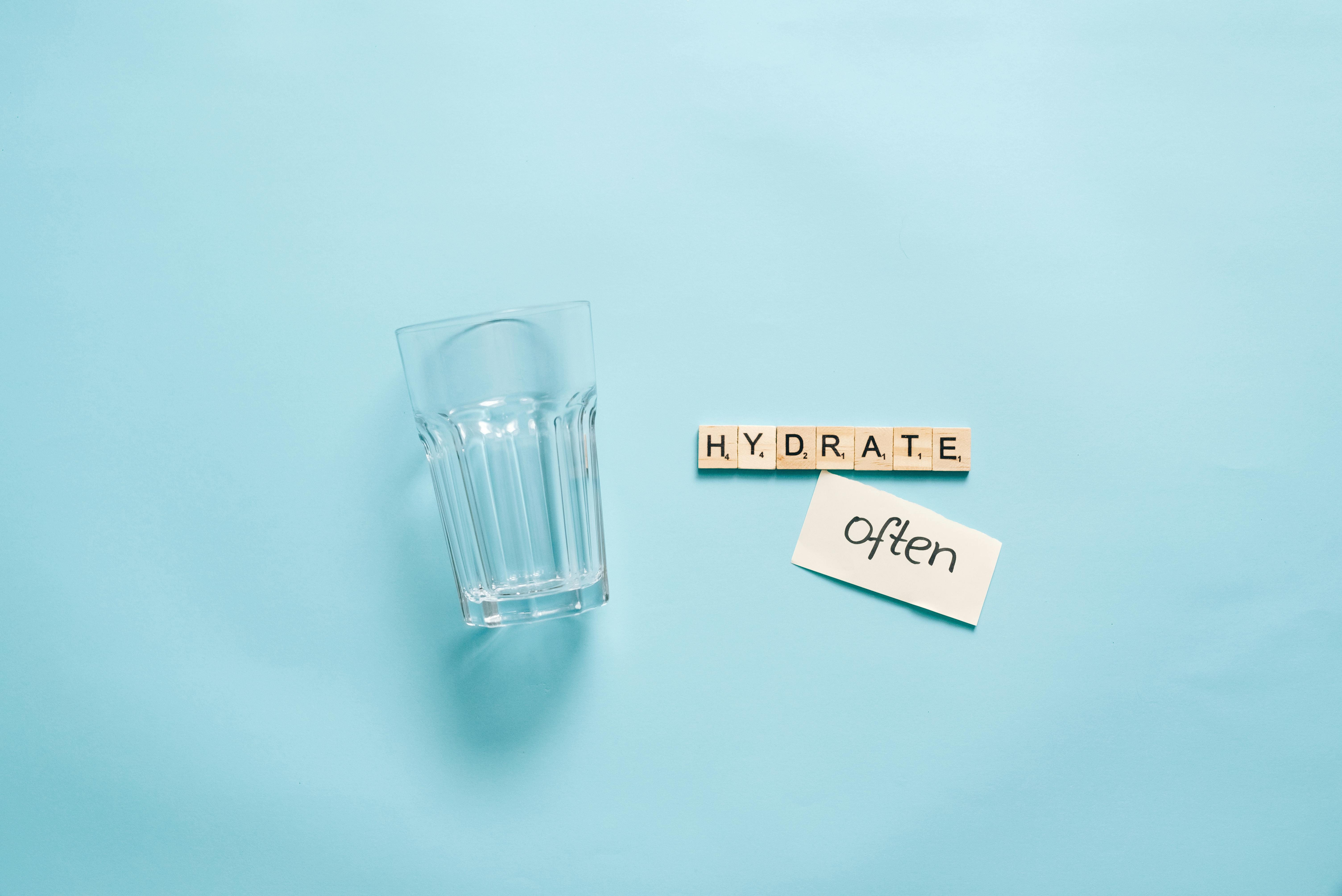 A minimalist composition of a glass with Scrabble tiles spelling 'HYDRATE' on a blue background.