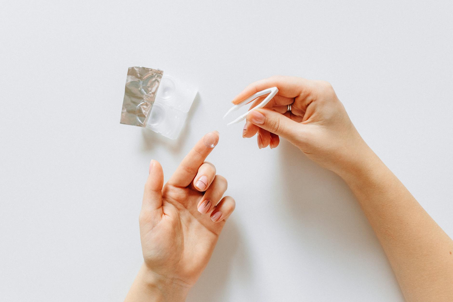 Hands holding a contact lens with tweezers next to a contact lens case on a white background.