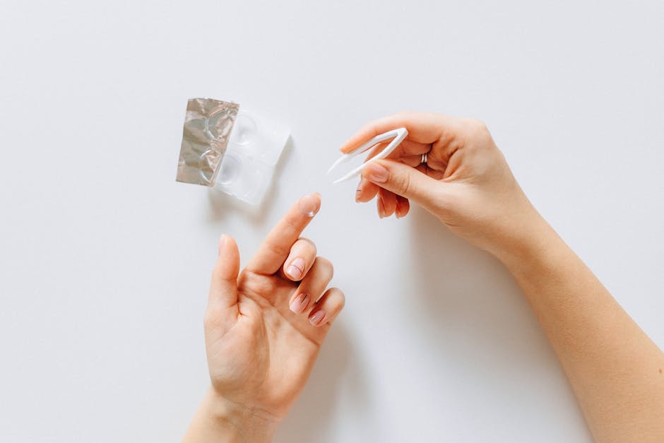 Hands holding a contact lens with tweezers next to a contact lens case on a white background.