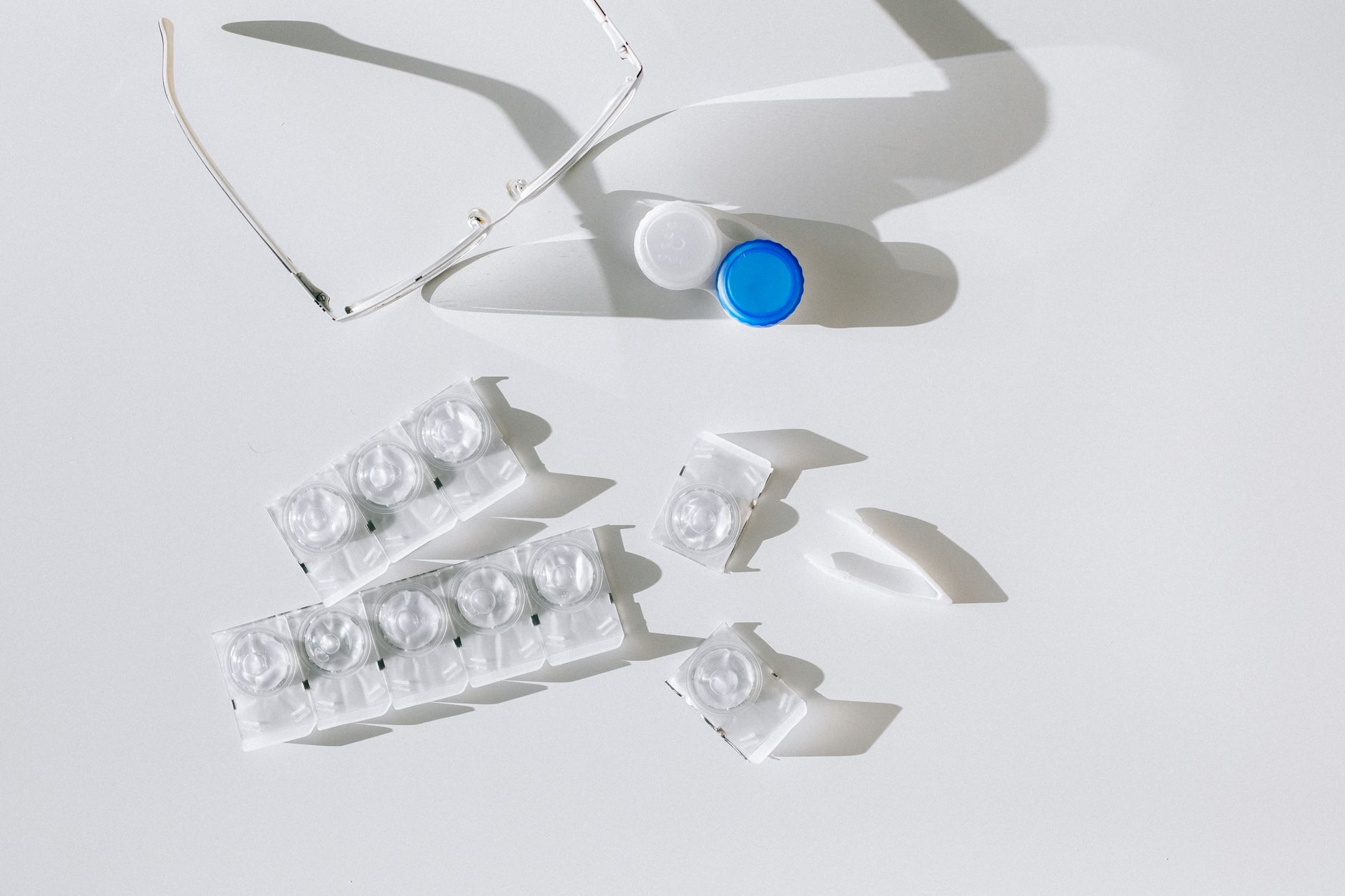 Flatlay of contact lens packages, case, and eyeglasses on a white surface with shadows.