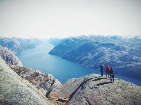 Three hikers stand atop a rocky peak, overlooking a breathtaking fjord landscape under a clear sky.