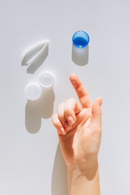 Close-up of a hand holding a contact lens with a container and solution nearby on a white surface.
