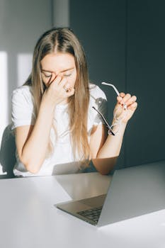 Young woman holds her glasses, experiencing digital eye strain in a bright office.
