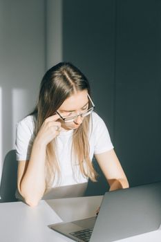 一位戴眼镜的年轻女子坐在桌旁，在阳光下使用笔记本电脑