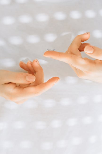 A detailed close-up of hands carefully handling a contact lens against a softly lit background.