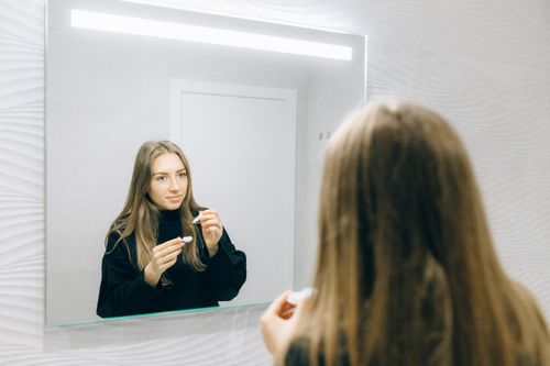 Free A young woman is seen applying a contact lens while looking in a mirror indoors. Stock Photo