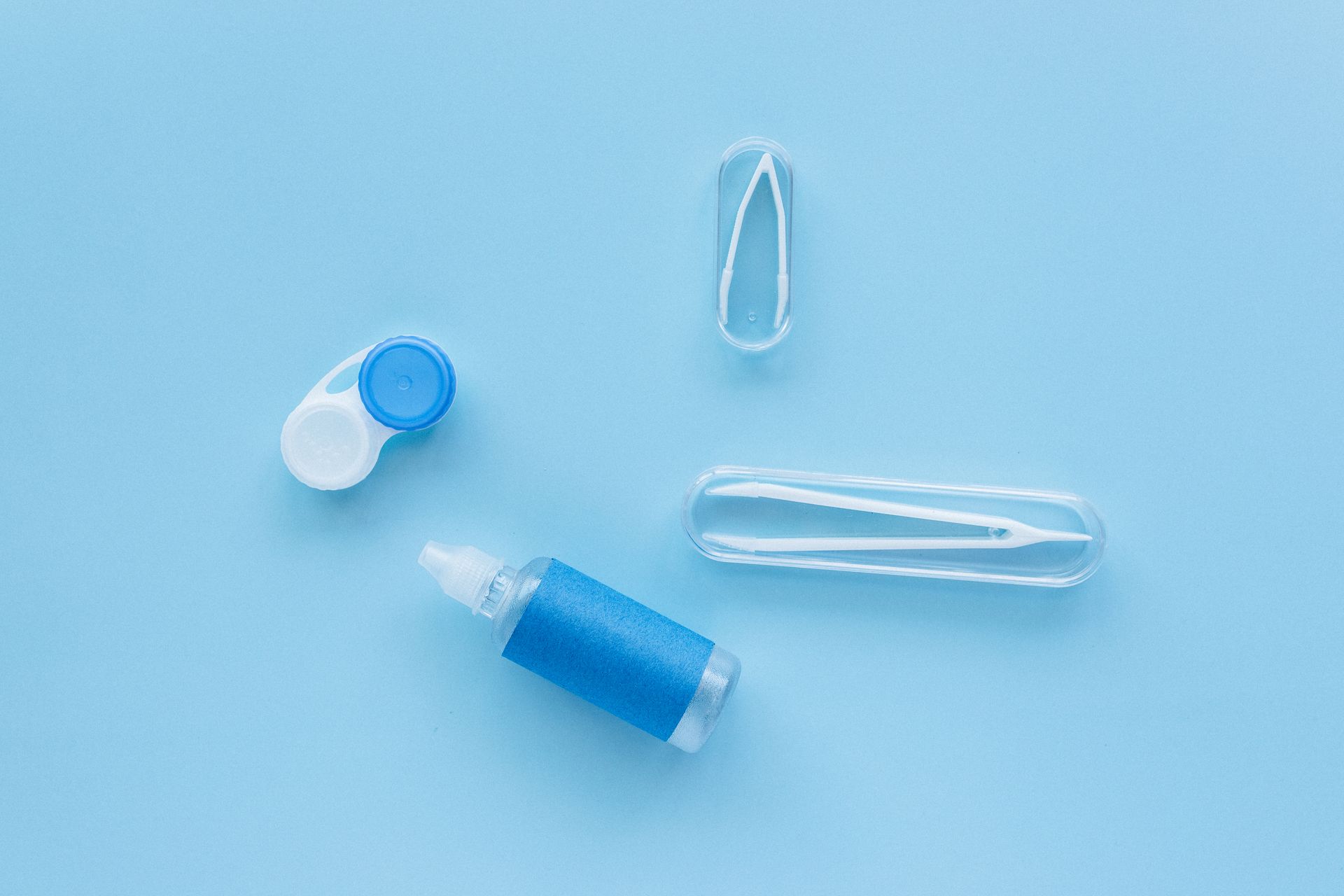 Flat lay of contact lens case, tweezers, and solution bottle on blue background.