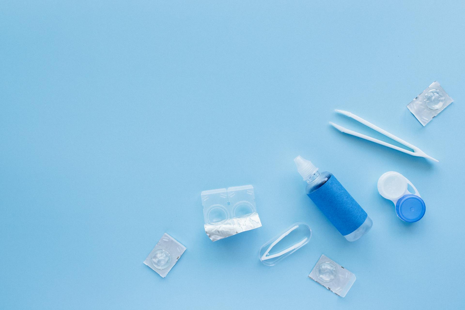 Flat lay of contact lens care kit with solution and tweezers on blue backdrop.