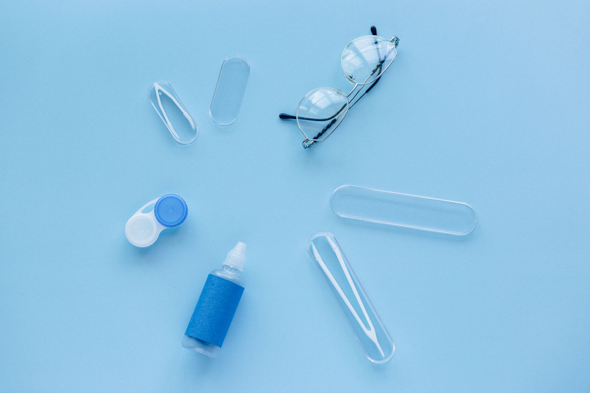 Flat lay of contact lens case, solution, and glasses on a blue background.