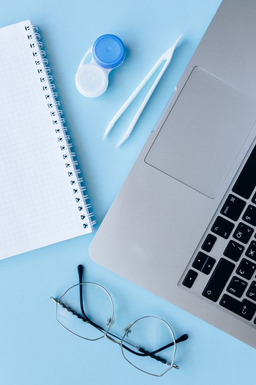 Free Minimalist flat lay of eyeglasses, a notebook, and a laptop on a blue background with contact lens case and tweezers. Stock Photo