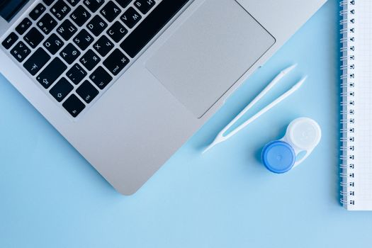 Top view of contact lens case, laptop, and notebook on a blue surface.