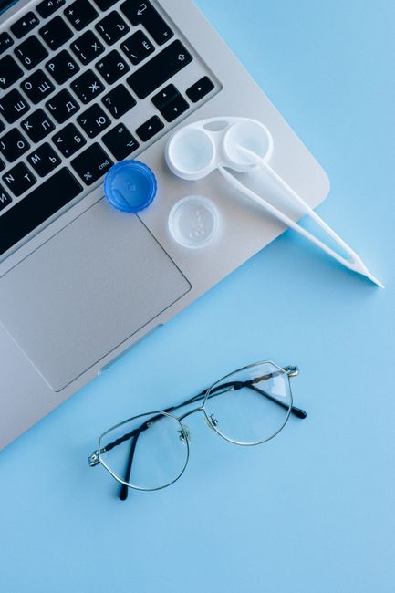 Flat lay of eyeglasses, contact lens case, and laptop on a blue surface.