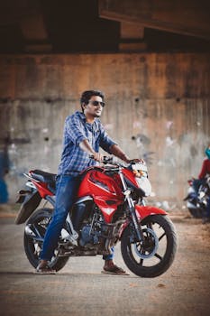 A man wearing sunglasses sits confidently on a red motorcycle under a bridge.