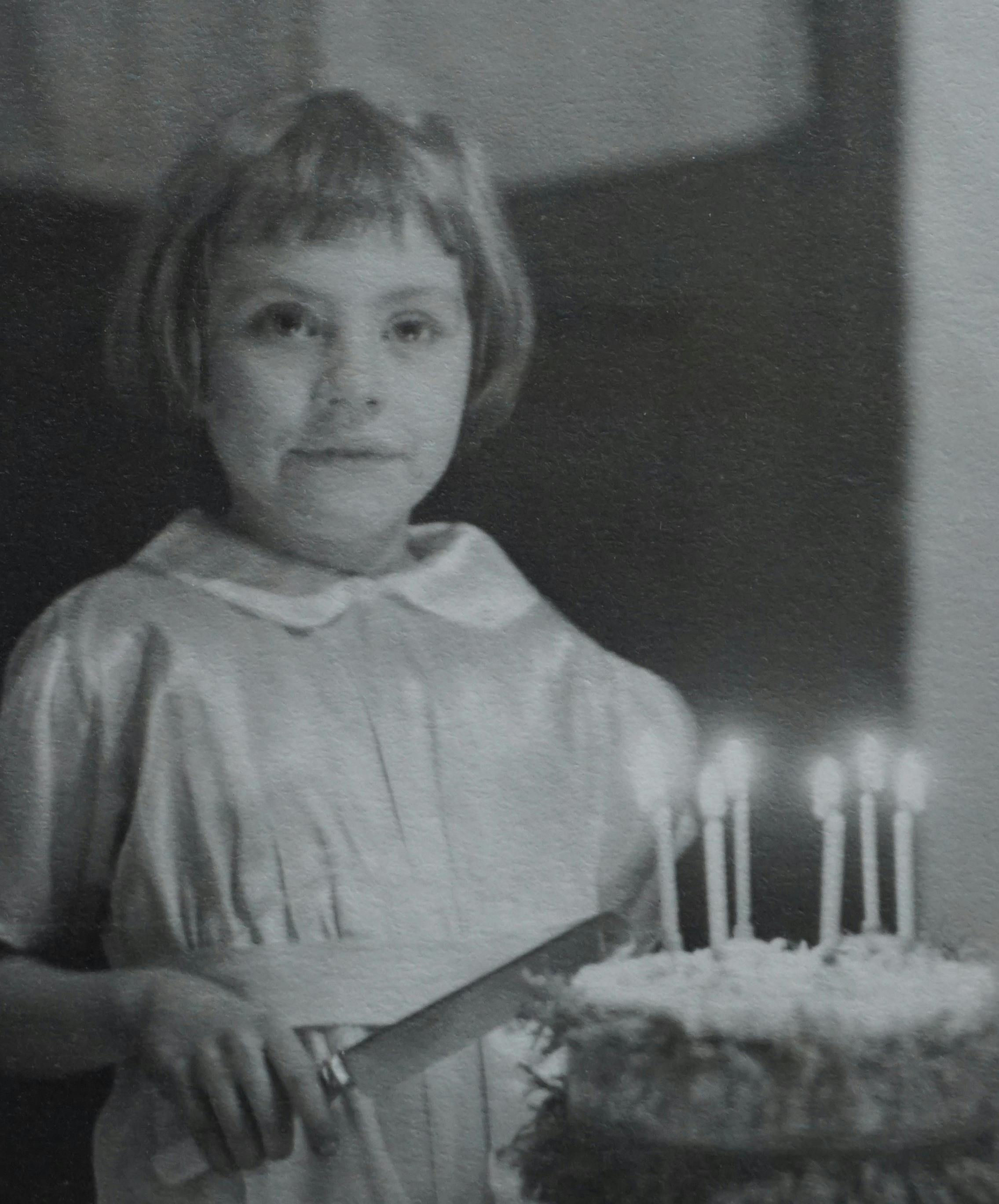 A young girl smiles by her birthday cake in a vintage grayscale photo, capturing a classic moment.