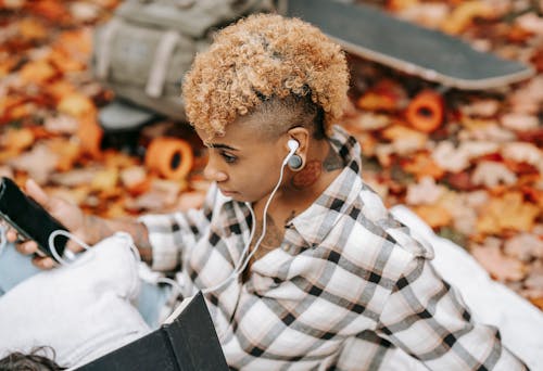 From above of African American female with earphones and smartphone sitting on blanket near skateboard and backpack on blurred background in autumn park