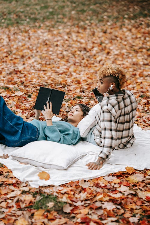 Side view of calm female reading book and lying on plaid on knees of African American friend listening to music while spending time in autumn park in daytime