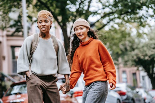 Free Smiling young black informal woman with backpack holding hands with cheerful Hispanic girlfriend while walking together along city street with trees Stock Photo