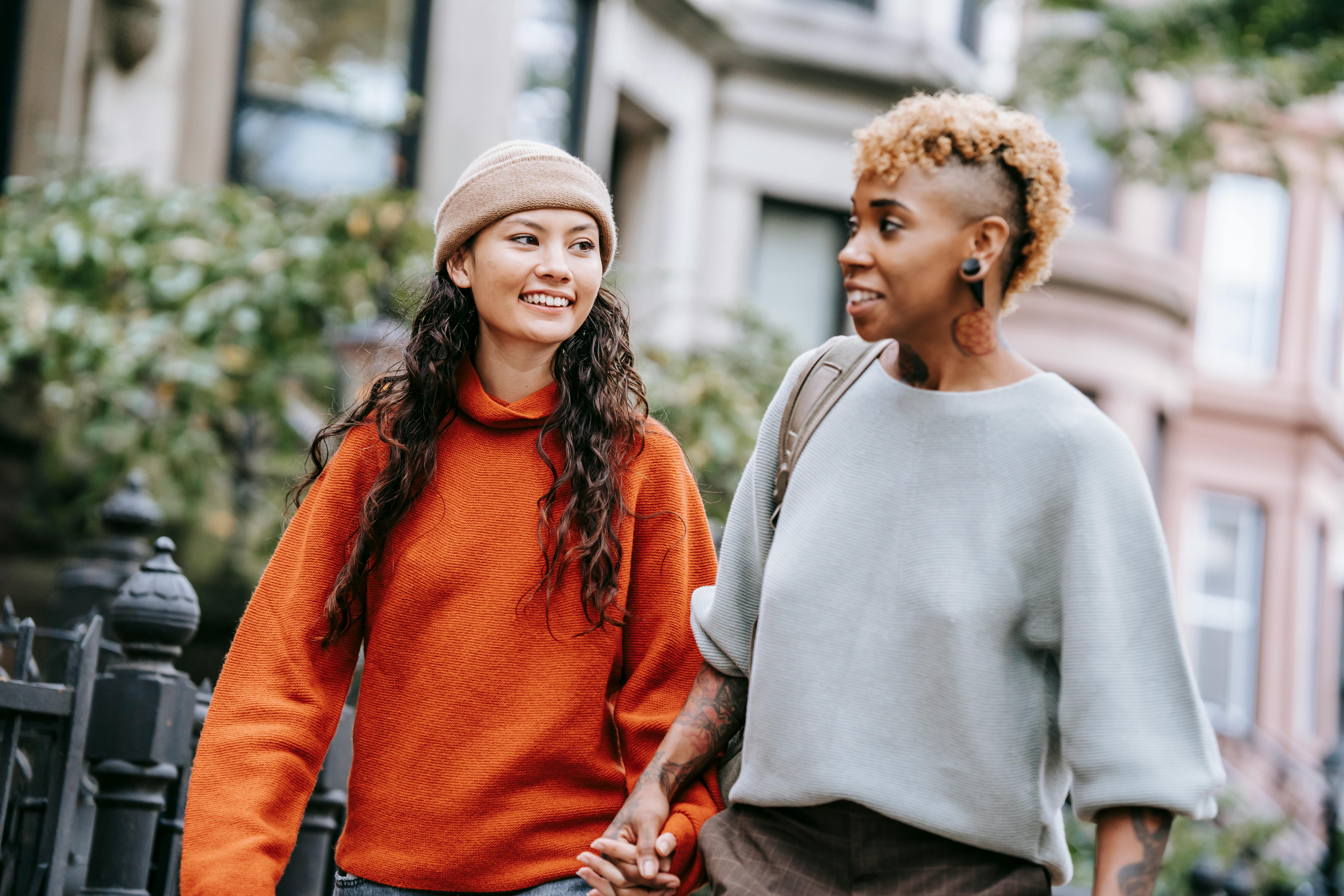 positive hispanic lady holding hands with girlfriend while walking near city buildings