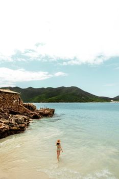 From above back view of anonymous female traveler in swimwear strolling on sandy ocean coast against mountain in summer