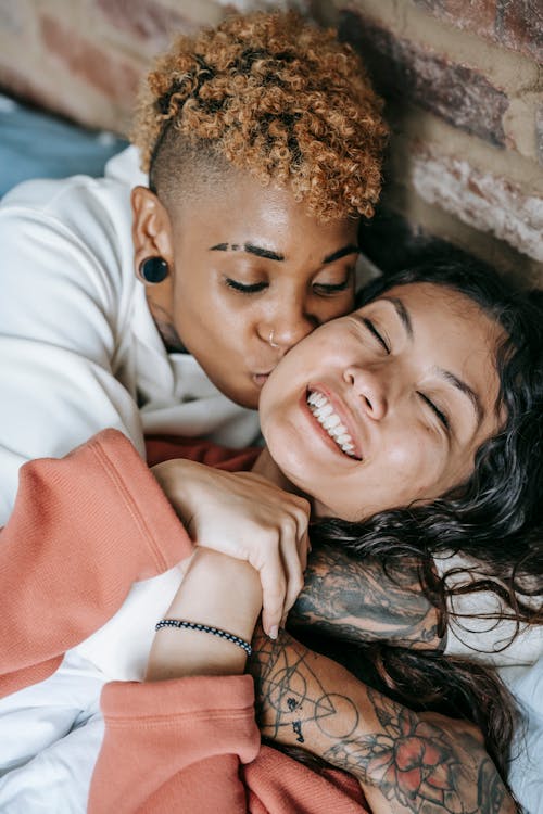 From above of happy young Hispanic woman lying on bed and embracing with African American informal girlfriend kissing gently cheek