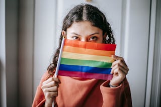 Young ethnic anonymous female demonstrating bright LGBT flag and looking at camera on blurred background