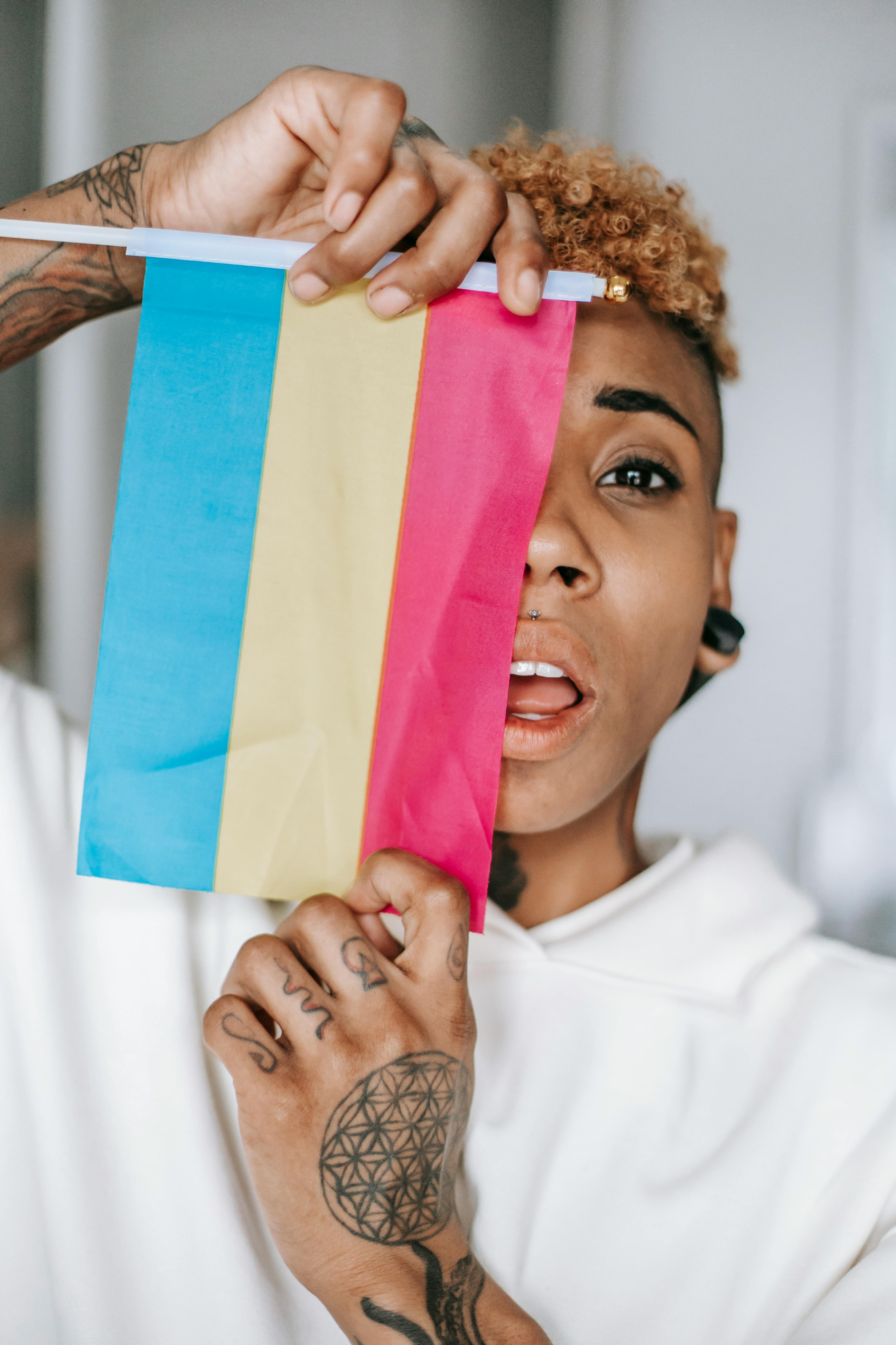 stylish black woman with tattoo hiding face behind pansexual pride flag