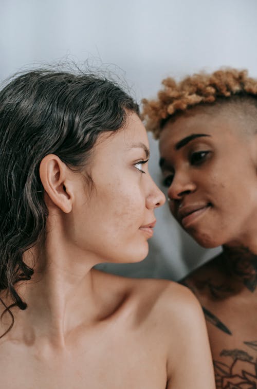 Free Young Hispanic woman with bare shoulders standing close to same sex African American girlfriend and looking on each other on white background Stock Photo