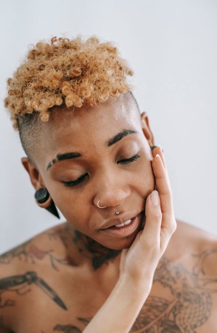 Anonymous woman touching face of African American female with tattoo and bare shoulder while standing on white background with closed eyes