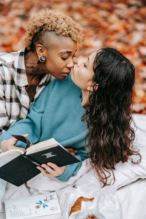 Free Side view of tender Hispanic woman with book kissing nose of African American girlfriend while lying on blanket during picnic in park on blurred background Stock Photo