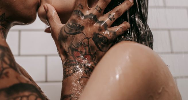 Side view of unrecognizable African American person with tattoo on hand kissing girlfriend with naked shoulders while standing under shower in bathroom with tiled wall