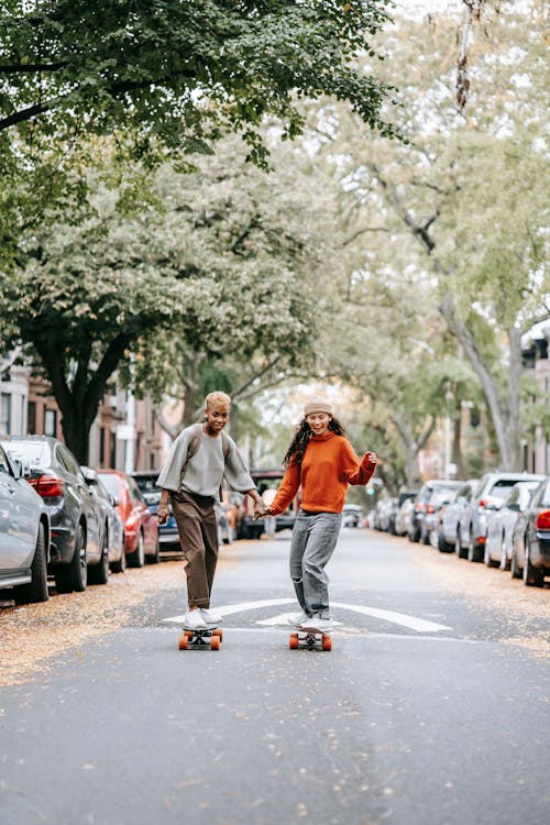 Free Happy Asian lady wearing warm sweater and hat holding hands with black lesbian partner while riding boards among parked cars in city street Stock Photo