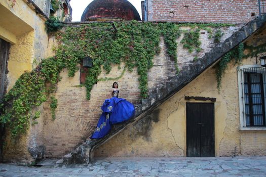 Woman in elegant blue gown gracefully posed on rustic concrete stairs with ivy-covered wall.