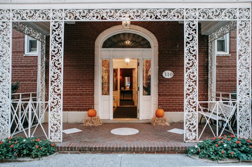 Exterior of residential brick building with doorway and white ornamental veranda with metal barriers located on street with blooming flowers