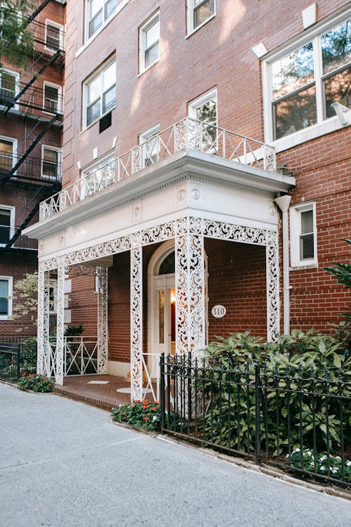 Exterior of residential brick house with white ornamental terrace located near metal fence with green plants in city on street