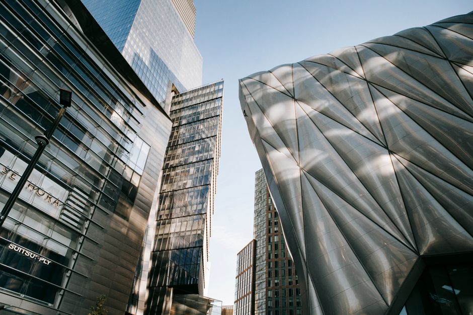 From below of contemporary office center with transparent walls located near futuristic building against blue sky in city on street
