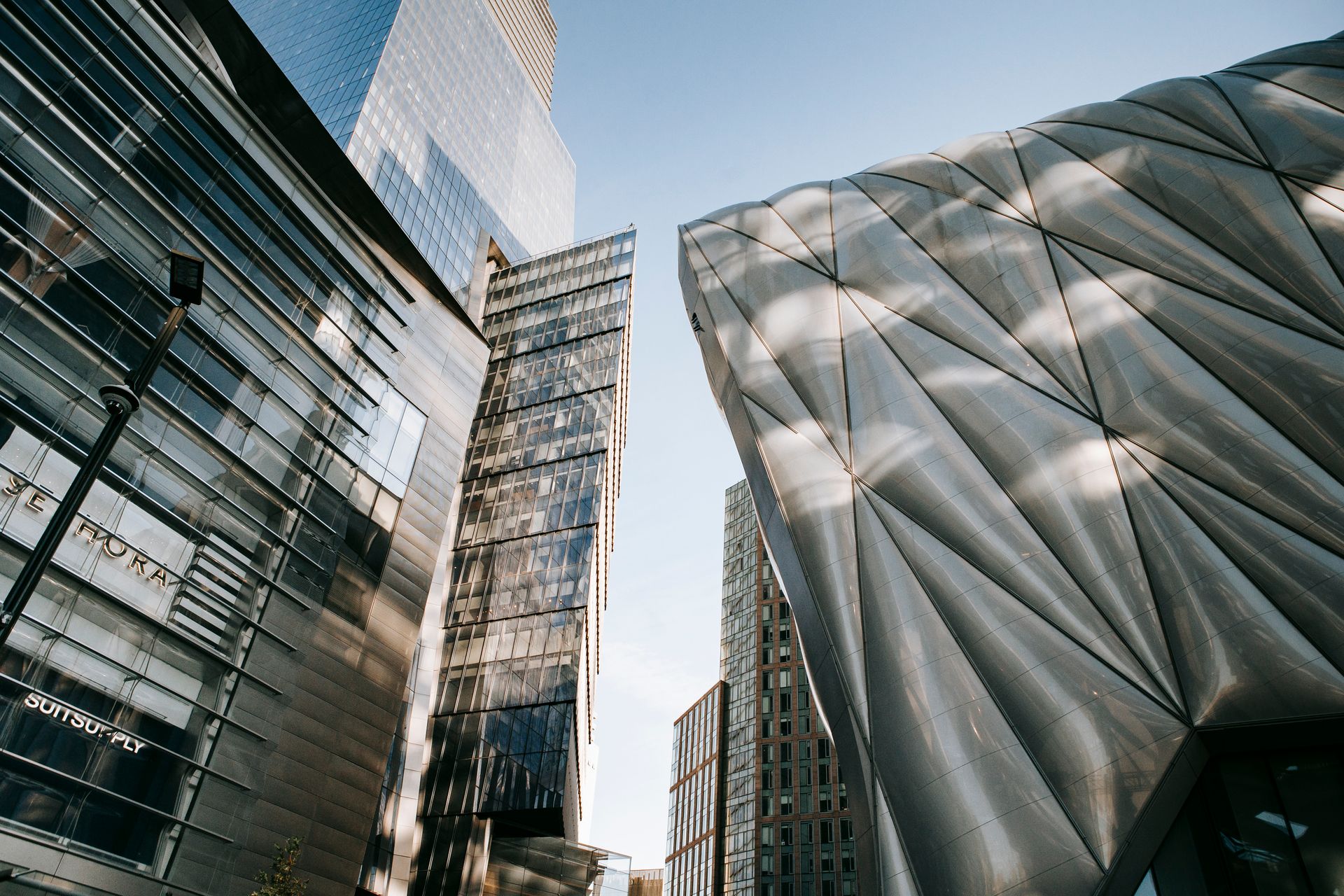 From below of contemporary office center with transparent walls located near futuristic building against blue sky in city on street