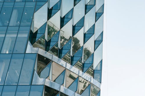 Corner of contemporary geometric building with reflection of cityscape on big glass transparent walls located on city street against cloudless sky