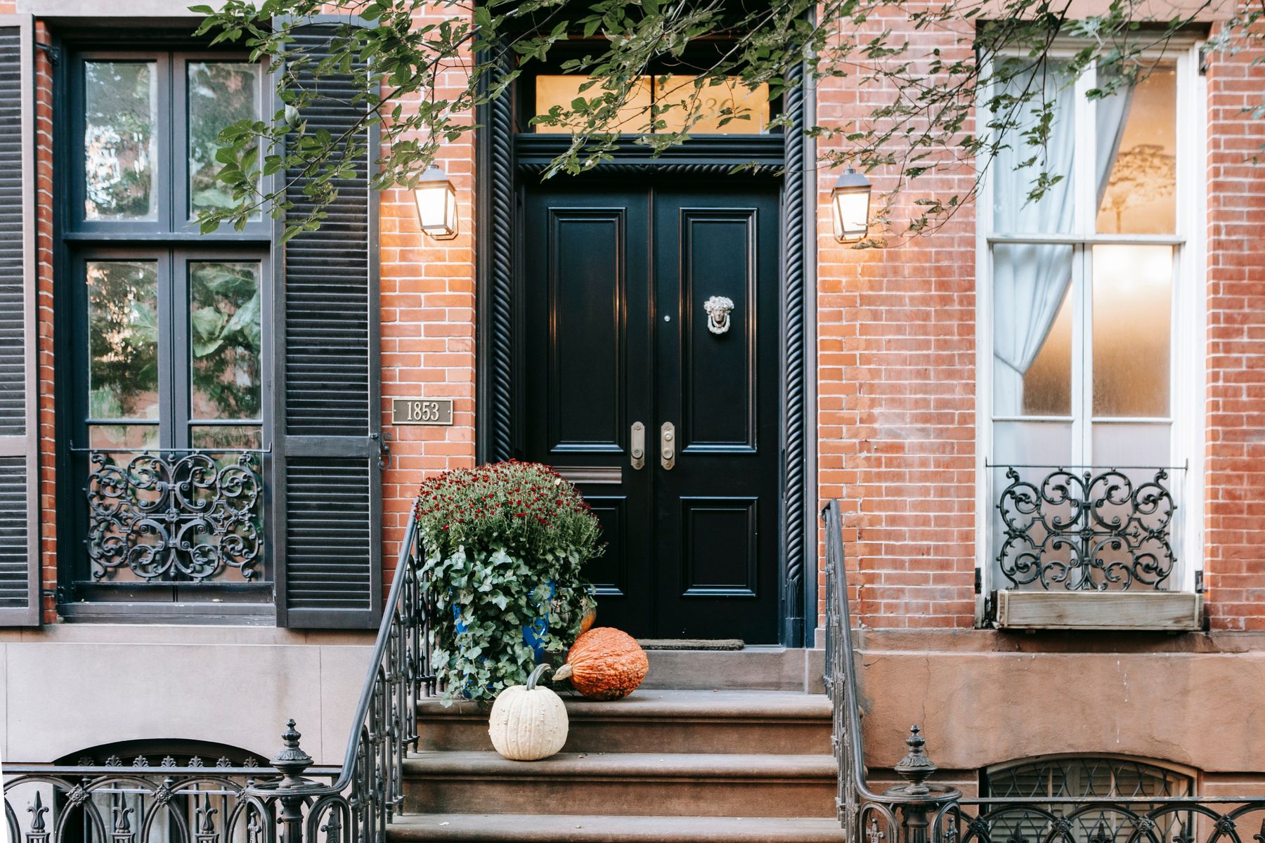 A black front door between brick walls and wrought-iron window grilles, with autumn plants on the stoop