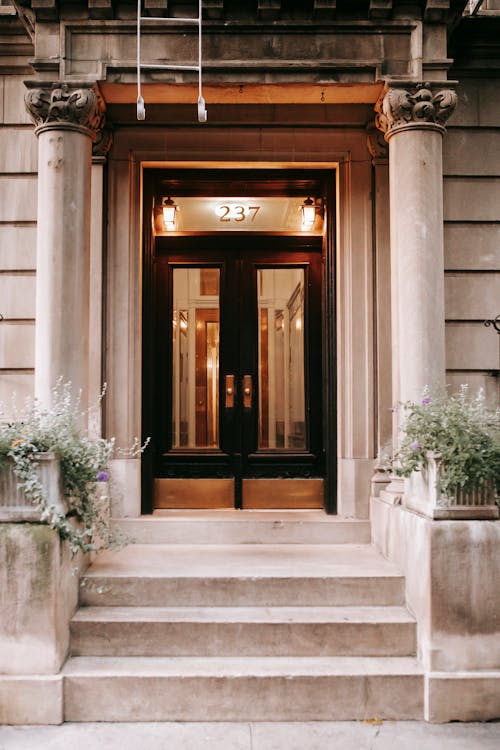 Exterior of aged entrance to apartment with pillars and flowers in pots and wooden door with glass at daytime